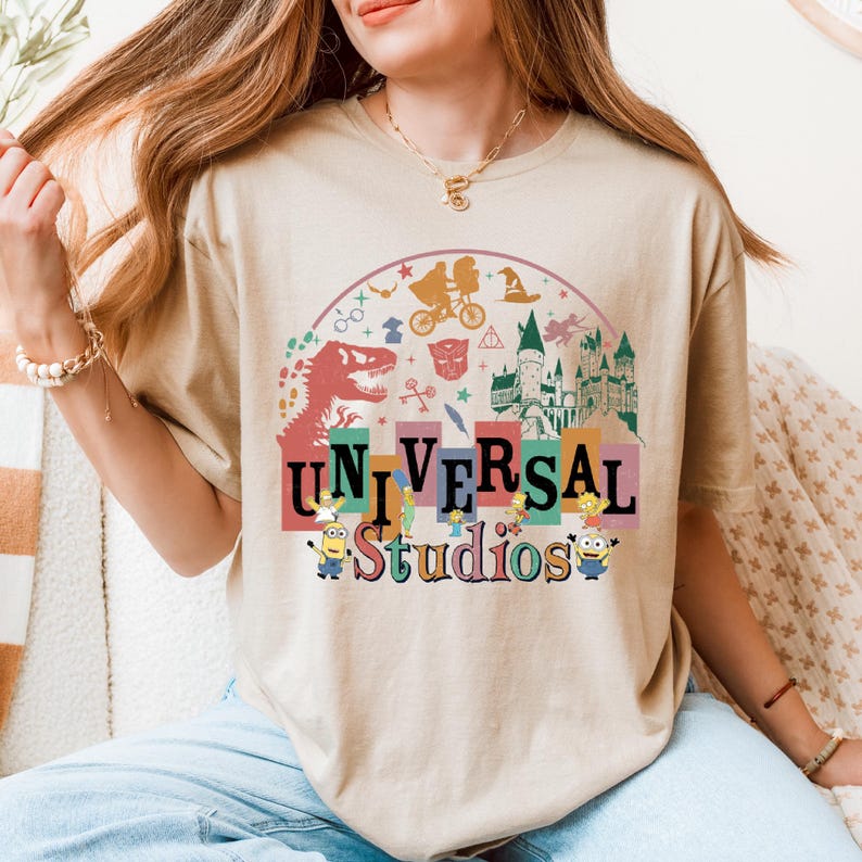 May include: Beige t-shirt with a vibrant "UNIVERSAL Studios" graphic. The design incorporates a dinosaur, Minions, and other theme park imagery. The shirt has a loose fit and is made of a soft material.