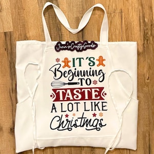 May include: White apron with pockets and long straps. The apron features the text "It's Beginning to Taste a Lot Like Christmas" in black and red, with gingerbread men and snowflakes. The apron also has the text "Jenn's Crafty Goods".