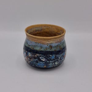 May include: A small, rounded ceramic cup featuring a smooth, tan-coloured rim that transitions into a mottled glaze of deep blue, light blue, and dark brown tones. The surface displays a textured, speckled finish with abstract dark markings.