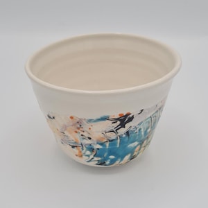 May include: A white ceramic bowl with a flared rim. The exterior has an abstract design with splashes of blue, orange, and black. The bowl is set against a plain white background.