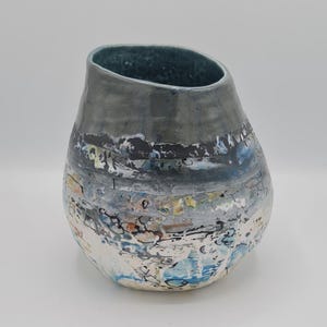 May include: A handmade ceramic vase with an unusual, asymmetrical form. The vase has a dark grey upper section and a lower part with a textured, multi-coloured design, including blue, white, and yellow accents. The interior is a deep teal colour.