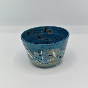 May include: A turquoise ceramic bowl with an irregular shape and abstract painted designs. The interior of the bowl has dark brown speckles. The bowl is a decorative item for the home. The bowl's measurements are in centimetres.