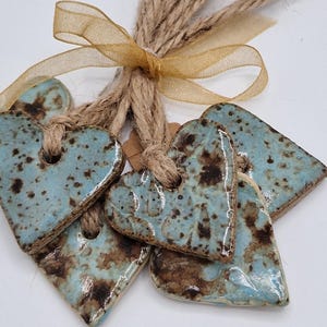 May include: A cluster of ceramic heart-shaped decorations in blue and brown hues, bound with twine and a gold ribbon. The ornaments feature a speckled glaze and are suitable for home decor. Each heart is approximately 5 cm wide.