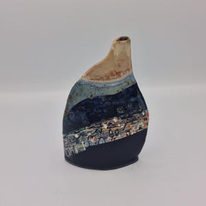 May include: A small ceramic vase with an asymmetrical, tapered neck. The surface features a layered glaze design with a beige top, a light blue middle section with dark speckles, and a solid matte black base separated by a decorative band.