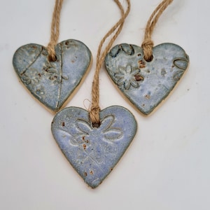 May include: Three blue ceramic heart decorations with floral patterns, each suspended by a natural twine cord. The hearts have a rustic, handmade appearance, ideal for home decor. Each heart is approximately 7 cm wide.