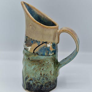 May include: A handmade ceramic jug with a textured surface. The jug features a tan rim and handle, with a blue interior. The body transitions from teal to dark blue, with brown and white accents. The jug has a unique, angled spout.