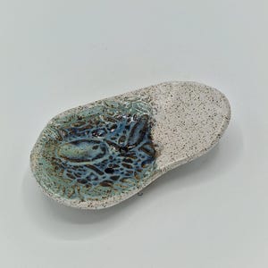 May include: An oval ceramic spoon rest with a speckled off-white exterior and interior rim. The centre features a textured, recessed pool of deep blue and turquoise glaze with a crackled, organic pattern that contrasts with the smooth speckled base.