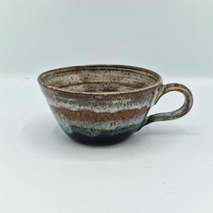 May include: A wide-rimmed ceramic mug featuring a speckled brown interior and a multi-coloured exterior glaze. The exterior displays horizontal bands of earthy brown, muted blue, and deep charcoal, with a curved handle attached to the side.
