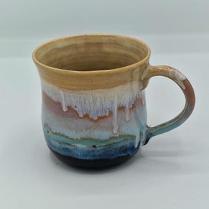 May include: A ceramic mug featuring a multi-coloured glaze with a tan rim that transitions into layers of white, pink, light blue, and dark blue. The glaze has a drip effect throughout the body, and the handle is coloured with shades of tan and blue.