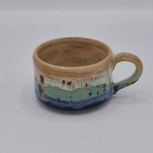 May include: A ceramic mug with a wide, low-profile shape and a single handle. The exterior features a layered glaze design transitioning from a tan rim to a light blue middle section, ending in a dark blue base with scattered brown speckles.