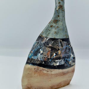 May include: A handcrafted ceramic vase with an unusual, asymmetrical form. The vase has a light blue top, a central section with abstract patterns in black, blue, and white, and a tan base. The vase is approximately 25 cm tall.
