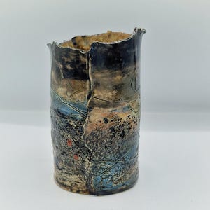 May include: A cylindrical ceramic vase with a textured surface. The vase features a colour palette of blues, blacks, and oranges. The rim is uneven, and the surface has a cracked appearance. The vase is suitable for home decor.