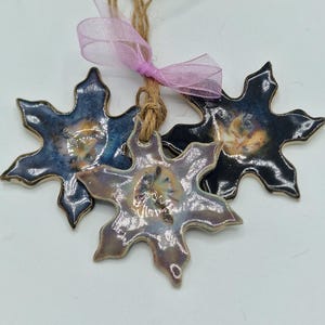 May include: Three ceramic snowflake ornaments with a glossy finish, featuring blue, black, and lavender hues with metallic centres. Each piece has a hole at the top, threaded with natural jute twine and tied together with a light pink ribbon.