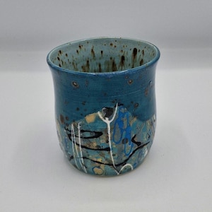 May include: A handcrafted ceramic tumbler in teal blue with a speckled interior. The exterior is decorated with abstract patterns in white, black, and yellow. The rim is angled, adding a unique touch to this handcrafted piece.