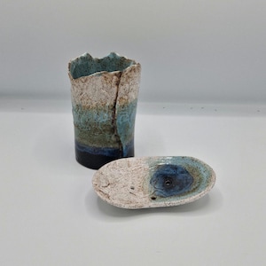 May include: A cylindrical ceramic vessel with a jagged, uneven rim and a textured, speckled glaze transitioning from white to blue and dark navy. Beside it sits an oval ceramic dish featuring a matching speckled finish and a central blue depression.