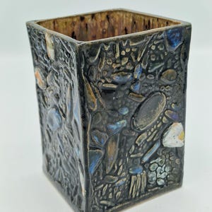 May include: A square ceramic vase with a textured, dark exterior. The surface is decorated with raised shapes and colours, including blue, brown, and white. The interior has a contrasting brown glaze.