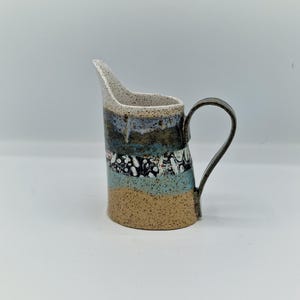 May include: A ceramic creamer jug featuring a speckled off-white rim, a dark brown and blue drip glaze, a central band of black and white mottled patterns, a light blue section, and a tan base with a dark brown curved handle on the side.