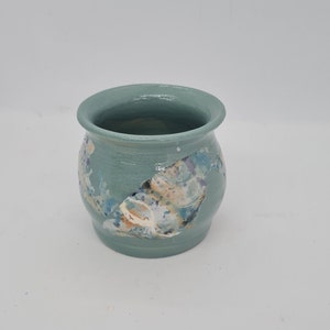 May include: A small, round, ceramic pot in a muted teal colour. The pot features a decorative, abstract design with splashes of white, blue, orange, and purple. The rim of the pot is slightly flared.