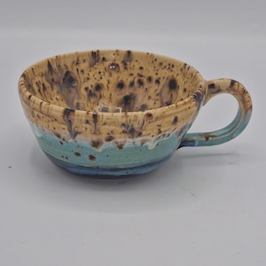 May include: A ceramic mug with a wide, shallow bowl shape and a single handle. The interior and upper exterior feature a cream-coloured glaze with dark brown speckles. The lower exterior is finished in a vibrant turquoise blue glaze.