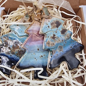 May include: Three ceramic Christmas tree ornaments with a glossy glaze in shades of blue, brown, and black. Each tree features a small star at the top, a jute twine loop for hanging, and a sheer ribbon tied at the base of the twine loop.