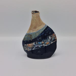 May include: A ceramic vase with an asymmetrical, organic shape. The upper portion is a light tan, transitioning into a band of mottled blue and green. A dark navy blue base features a horizontal textured strip with flecks of orange and white.