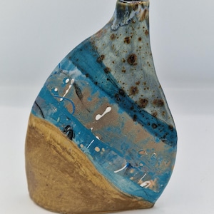 May include: A ceramic bud vase with an asymmetrical, teardrop-like silhouette. The surface features a textured, sandy-brown base transitioning into a vibrant blue glaze, accented with dark brown speckles and small white paint-like splatters.
