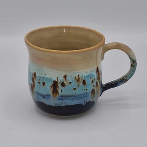 May include: A ceramic mug featuring a rounded body and a curved handle. The upper portion is a smooth beige, transitioning into a vibrant blue middle section with brown speckled accents, and finishing with a solid dark navy blue base.
