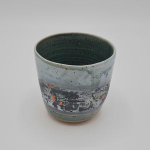 May include: A handcrafted ceramic cup with a distinctive design. The cup has a dark green interior and a light blue exterior with a textured, abstract pattern in black, grey, and orange. The cup is approximately 10 cm tall.