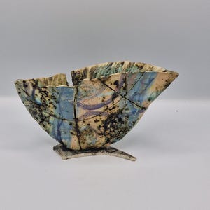 May include: A hand-built ceramic bowl with an irregular, flared rim and a textured surface. The piece features a mottled glaze in shades of blue, cream, and brown, with dark, incised geometric lines and a rough, weathered appearance on a base.
