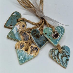 May include: A collection of six ceramic heart-shaped ornaments with glossy glazes in shades of blue, turquoise, and cream, featuring speckled and marbled textures. Each heart is suspended by a brown jute twine and tied with a sheer white ribbon.
