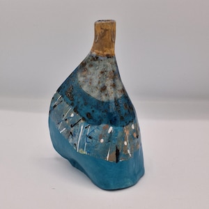 May include: A small, asymmetrical ceramic vase with a narrow neck. The surface features a layered design with a tan neck, a speckled blue and white midsection, and a solid turquoise base, accented by thin vertical white lines and dark markings.