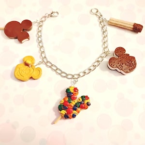 May include: Silver chain bracelet with five charms. Charms include a brown Mickey Mouse-shaped popsicle, a yellow Mickey Mouse-shaped cookie, a rainbow-colored Mickey Mouse-shaped treat, a brown ice cream sandwich, and a brown churro.
