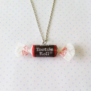 May include: A silver chain necklace with a Tootsie Roll candy charm. The candy is wrapped in red and white paper and has the words "Tootsie Roll" printed on it.
