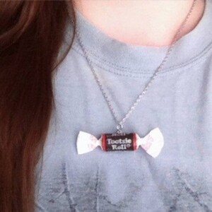 May include: A silver chain necklace with a Tootsie Roll candy charm. The charm is a brown and red Tootsie Roll with the words "Tootsie Roll" printed on it.
