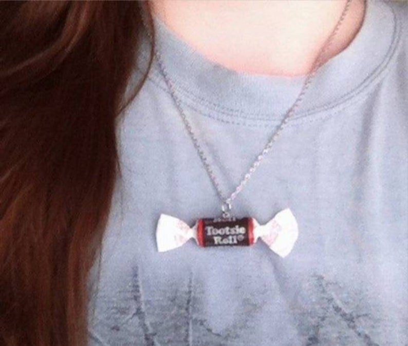 May include: A silver chain necklace with a Tootsie Roll candy charm. The charm is a brown and red Tootsie Roll with the words "Tootsie Roll" printed on it.