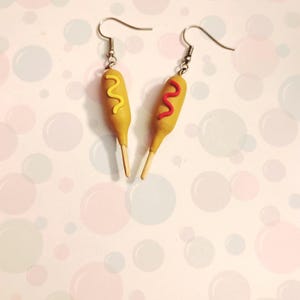 May include: A pair of novelty earrings shaped like miniature corn dogs. Each earring features a yellow corn dog with a wooden stick, and a squiggle of red or yellow topping. The earrings have silver-colored fish hook closures.
