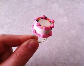 May include: A pink and white cake ring with colorful sprinkles. The ring is made of polymer clay and is a fun and unique piece of jewelry.