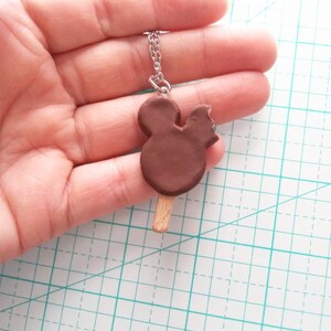 May include: A brown chocolate Mickey Mouse ice cream bar charm on a silver chain necklace. The charm is a bit melted and has a bite taken out of it.