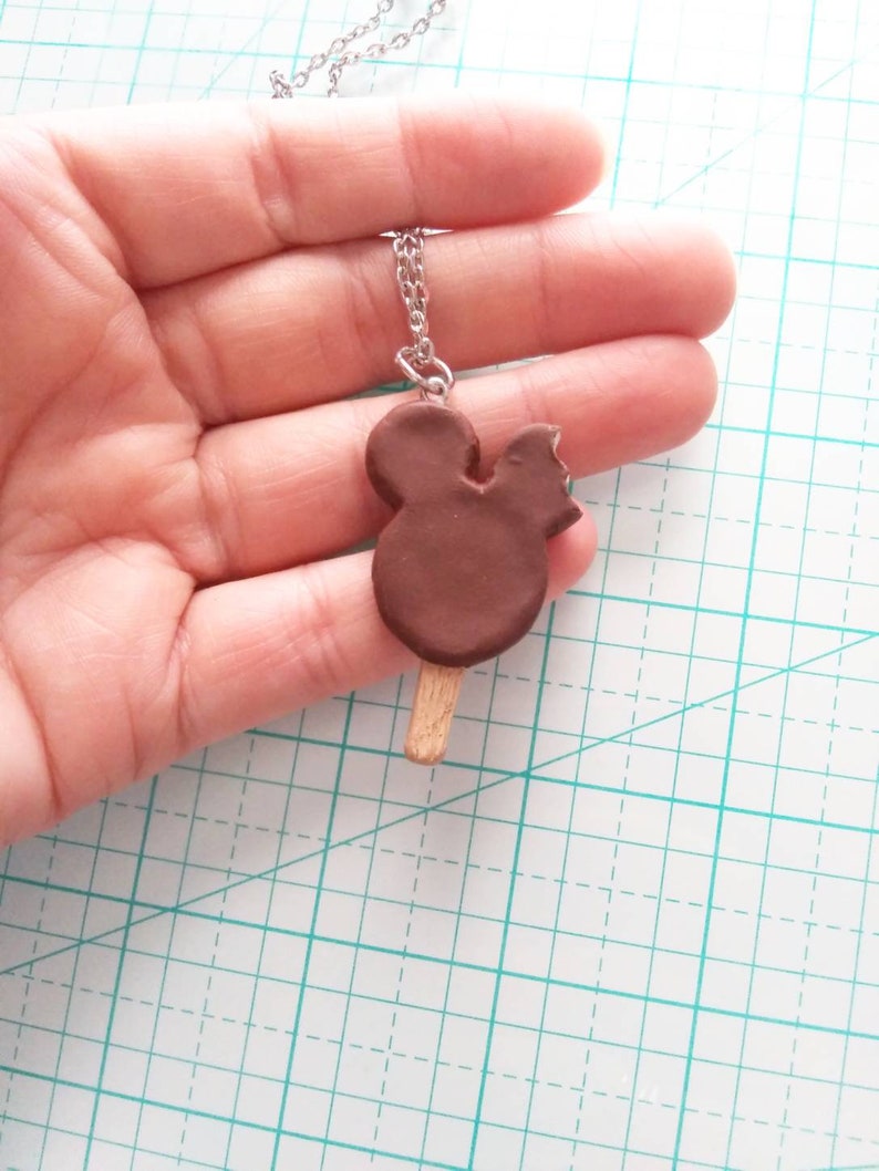 May include: A brown chocolate Mickey Mouse ice cream bar charm on a silver chain necklace. The charm is a bit melted and has a bite taken out of it.