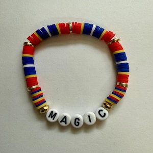 Puede incluir: Una pulsera colorida con cuentas de disco rojas, azules, amarillas y blancas. La palabra "MAGIC" está escrita con cuentas de letras blancas. Cuentas de metal doradas separan secciones de la pulsera.