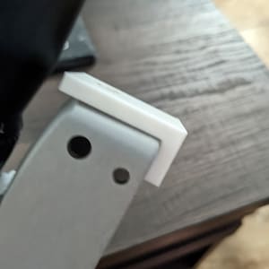 May include: A close-up of a white plastic piece attached to a silver metal object. The plastic piece has a rectangular shape with rounded edges.