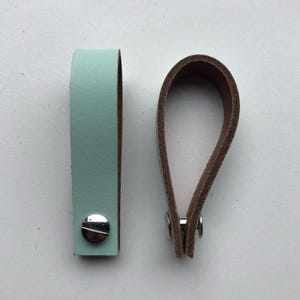 Op de afbeelding: Twee leren sleutelhangers. Eén is lichtblauwgroen van kleur met een zilveren klinknagel. De andere is een lus van bruin leer. De sleutelhangers zijn ontworpen om sleutels vast te houden.