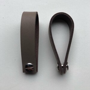 Op de afbeelding: Twee leren lussen in bruintinten. Eén is een lichtere taupe kleur, bevestigd met een zilveren schroef. De andere is donkerbruin, vastgezet met zilveren klinknagels. Beide zijn ontworpen voor handvatten of decoratieve doeleinden.