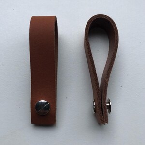 Op de afbeelding: Twee bruine leren treklussen met zilverkleurige klinknagels. Eén lus is plat, de andere is gelust. Het leer heeft een gladde textuur. Deze lussen zijn waarschijnlijk voor meubels of andere decoratieve doeleinden.