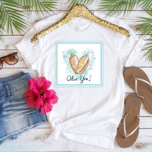Peut inclure: T-shirt blanc avec un graphique carré bleu et vert représentant deux coquillages en forme de cœur. Le texte "Olive You!" est écrit sous les coquillages.