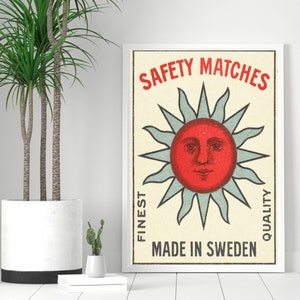 May include: A framed vintage-style poster with a red sun face design and the words "SAFETY MATCHES" in red. The poster also includes the text "FINEST QUALITY" and "MADE IN SWEDEN". A large potted plant is to the left of the poster.
