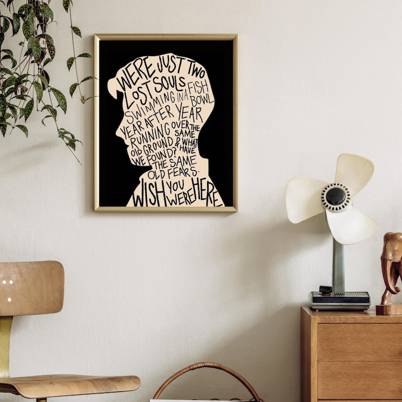 May include: A black and white print of a silhouette of a person's head with the words "WERE JUST TWO LOST SOULS FISH SWIMMING IN A BOWL YEAR AFTER YEAR RUNNING OVER THE OLD GROUND & WHAT WE FOUND? HAVE THE SAME OLD FEARS. WISH YOU WERE HERE" inside the silhouette.