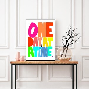 May include: A framed print with the text "ONE DAY AT A TIME" in colorful, bold letters on a white background. The print is displayed on a wooden table with a vase of branches.