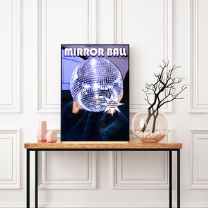 May include: A poster with the text "MIRROR BALL" in a retro font. A disco ball is in the center of the poster, partially obscured by a person wearing a blue fur coat. The text "I'LL SHOW YOU A REFLECTION OF YOURSELF TONIGHT" is in a white starburst shape.