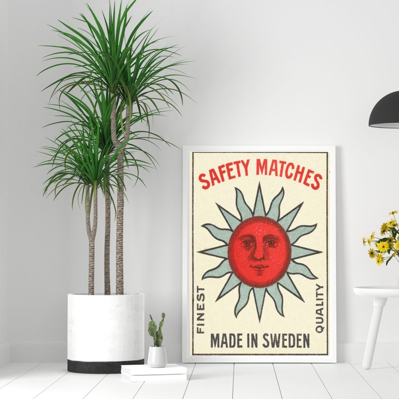 May include: A framed vintage-style poster with a red sun face design and the words "SAFETY MATCHES" in red. The poster also includes the text "FINEST QUALITY" and "MADE IN SWEDEN". A large potted plant is to the left of the poster.